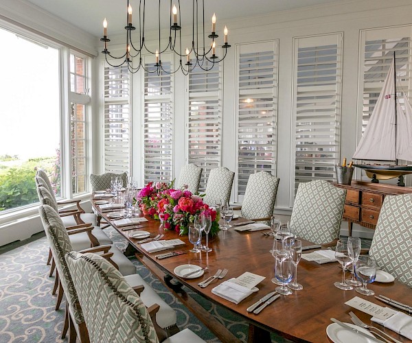 A dining room with a long table set for a meal, featuring elegant chairs, floral centerpiece, chandelier, and a model sailboat decor.