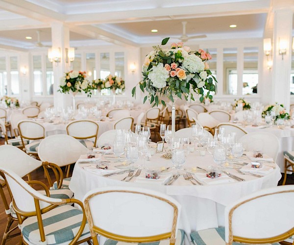 An elegantly decorated banquet hall with round tables, floral centerpieces, glassware, and neatly arranged place settings.