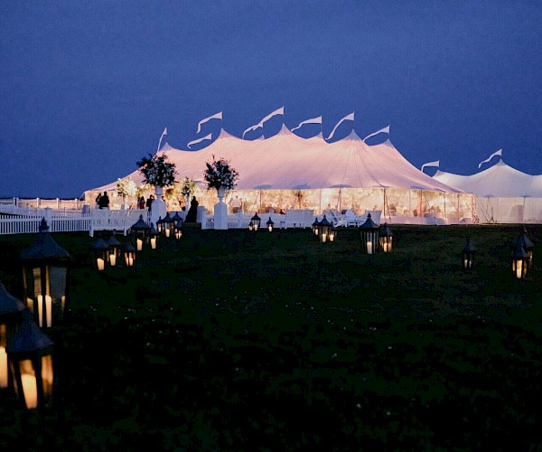 An illuminated tent at dusk with glowing lanterns lining a path, set against a dark sky, suggesting a festive outdoor event.