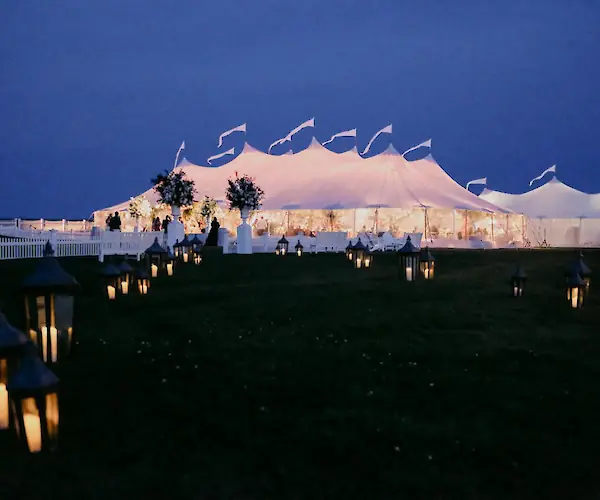 An illuminated tent at dusk with glowing lanterns lining a path, set against a dark sky, suggesting a festive outdoor event.