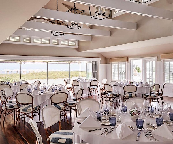 The image shows a banquet hall with round tables elegantly set for an event, featuring glassware, white napkins, and scenic outdoor views.