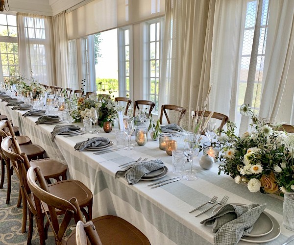 A long dining table is elegantly set with folded napkins, plates, glasses, candles, and floral arrangements in a bright room.