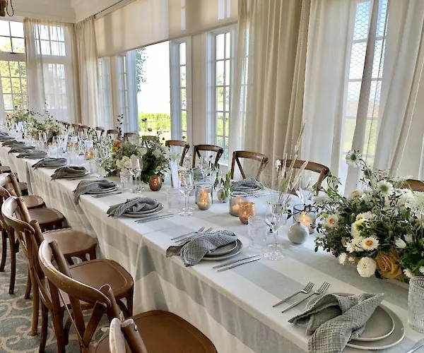 A long dining table is elegantly set with folded napkins, plates, glasses, candles, and floral arrangements in a bright room.