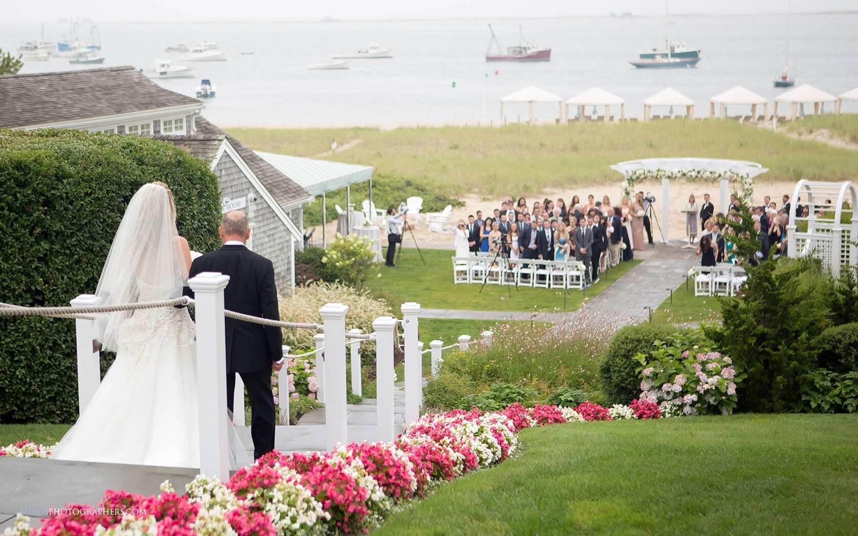 A bride is walking down a flower-lined path towards a seated wedding gathering by the sea under a cloudy sky.