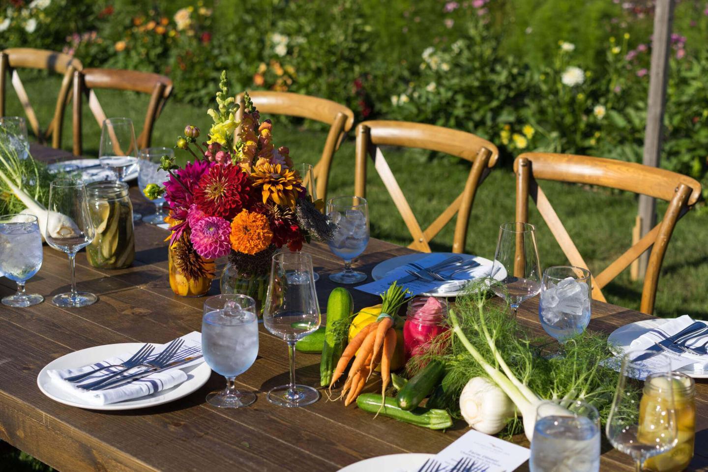 A beautifully set outdoor table with colorful flowers, fresh vegetables, glassware, and napkins, surrounded by chairs in a garden setting.