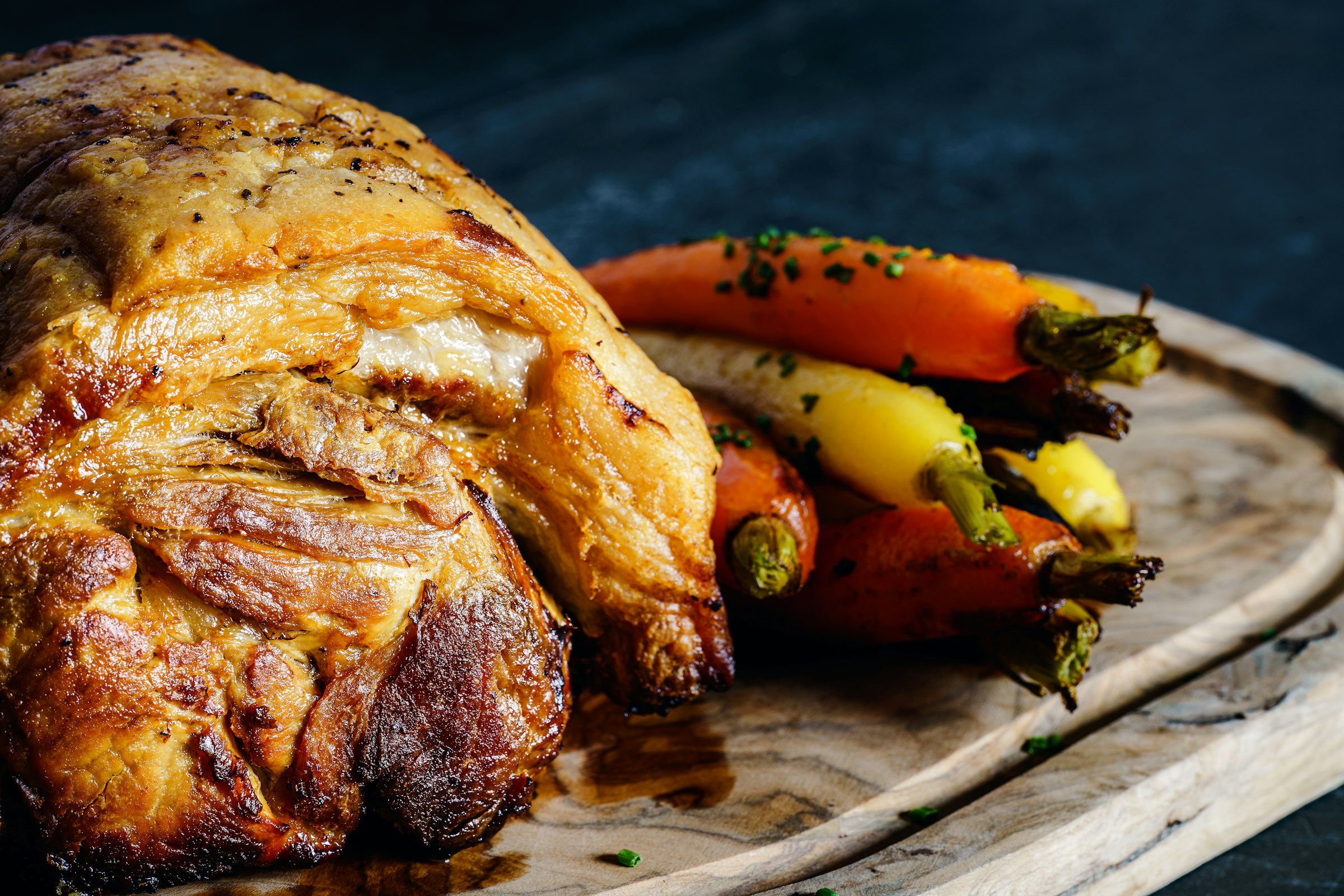 A roasted piece of meat with crispy skin sits next to colorful roasted carrots on a wooden board.