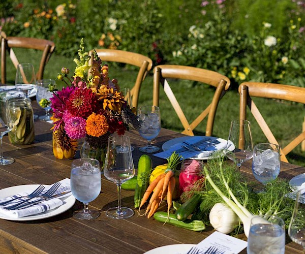 A beautifully set outdoor table with colorful flowers, fresh vegetables, and elegant tableware under a sunny sky.