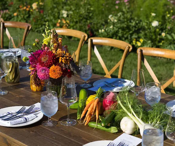 A beautifully set outdoor table with colorful flowers, fresh vegetables, and elegant tableware under a sunny sky.