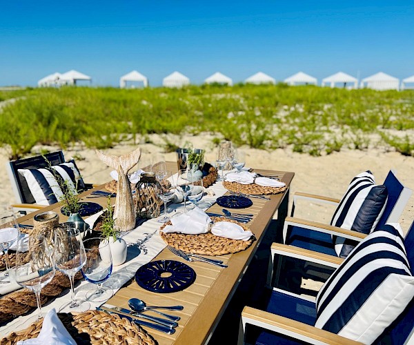 A beautifully set outdoor dining table on a sandy beach with striped cushions, surrounded by greenery and distant white tents.