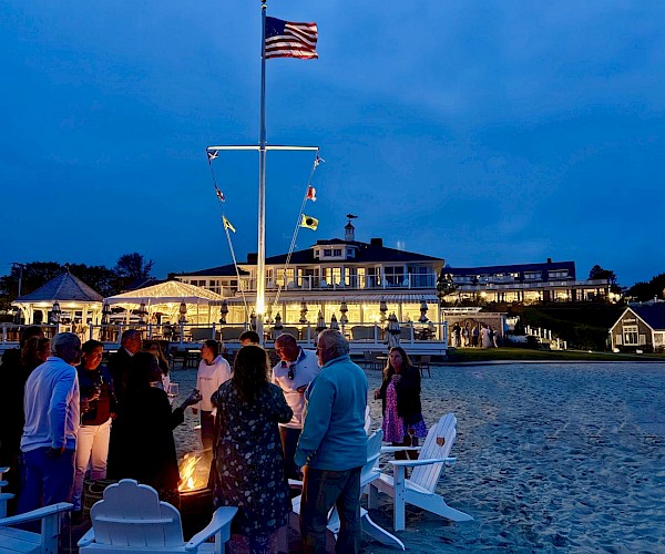 A group of people stand around a fire pit on a beach. There's a building in the background with warm lights.