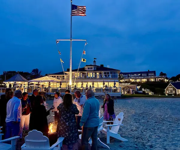 A group of people stand around a fire pit on a beach. There's a building in the background with warm lights.