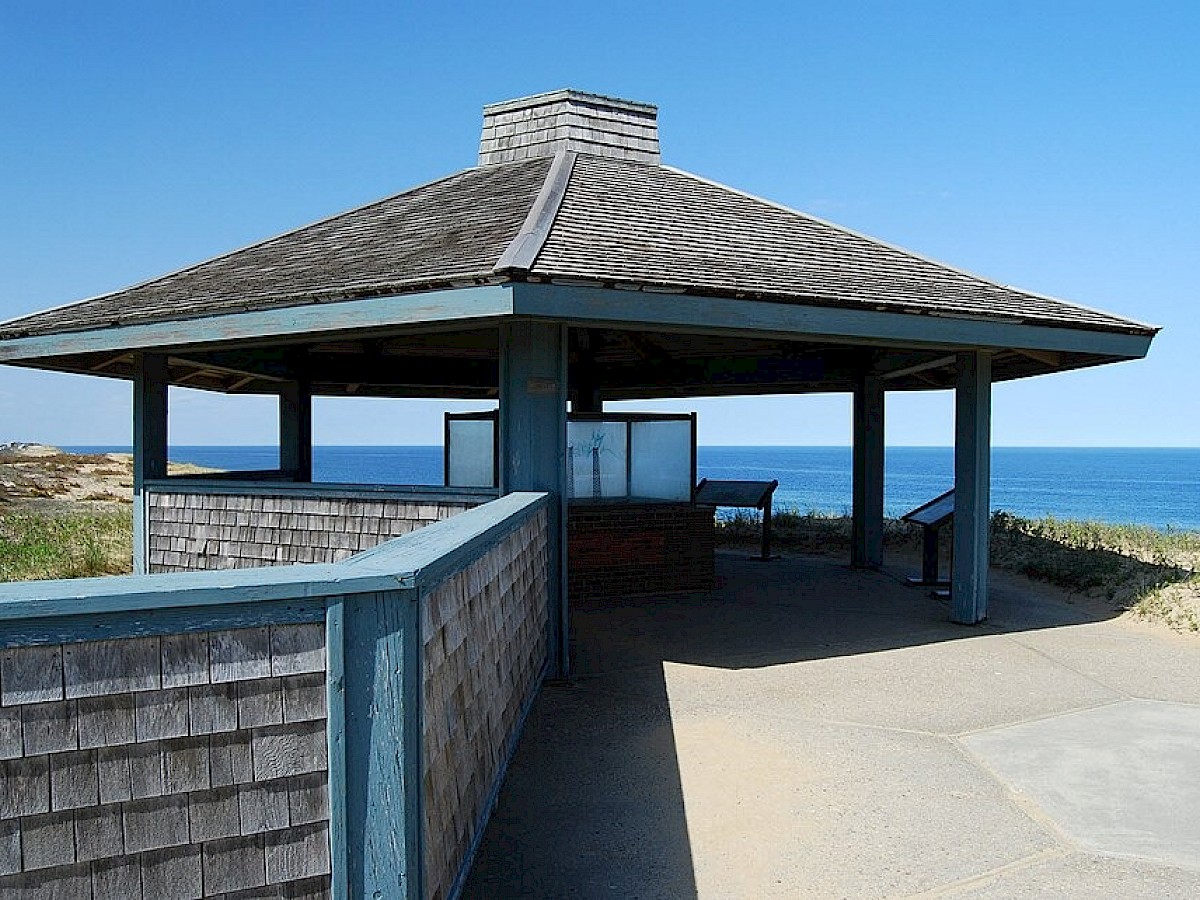 A wooden pavilion overlooking the ocean with informational signs, situated on a coastline on a clear day.