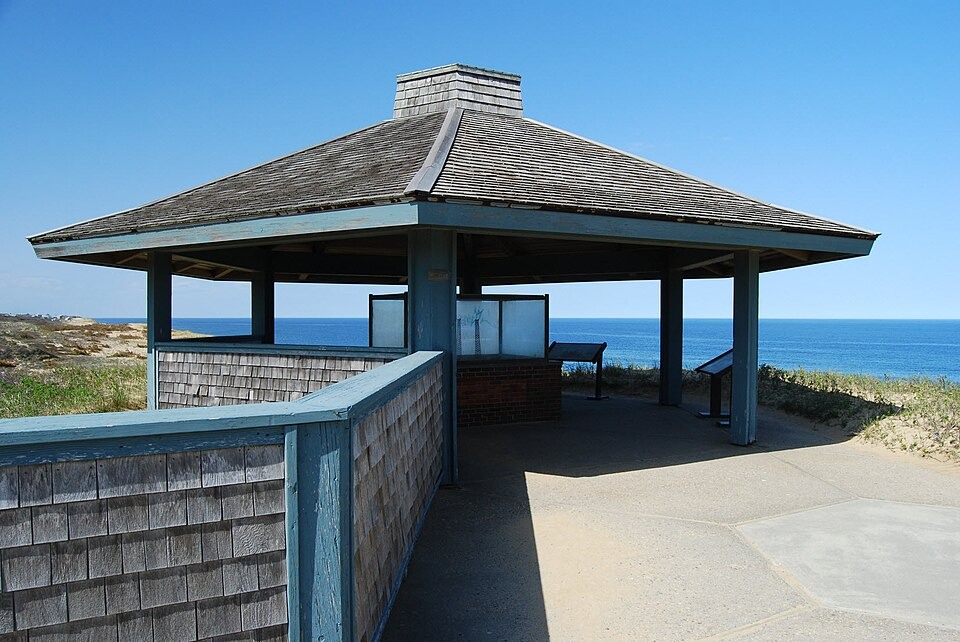 A wooden pavilion overlooking the ocean with informational signs, situated on a coastline on a clear day.