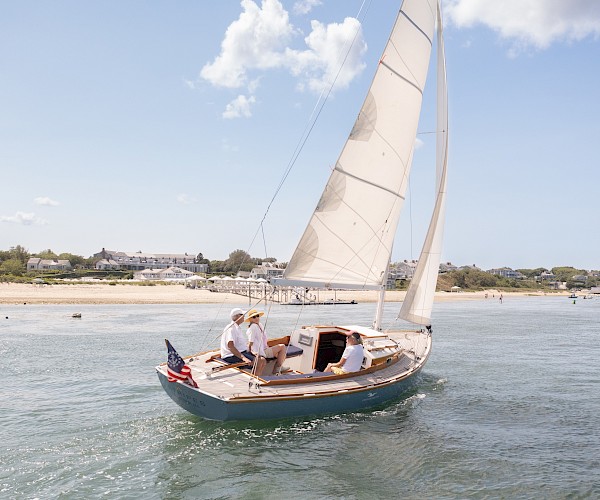 A sailboat with three people on board navigates near a sandy coastline with buildings in the background and a clear, blue sky overhead.