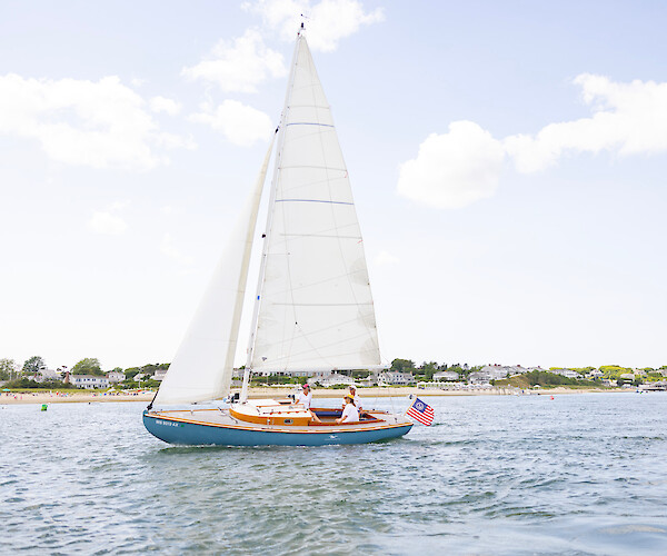 A sailboat with three people on board navigates near a sandy coastline with buildings in the background and a clear, blue sky overhead.