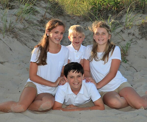 A group of four children in white shirts and shorts pose on a sandy beach, surrounded by grass.