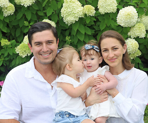 A group of four children in white shirts and shorts pose on a sandy beach, surrounded by grass.