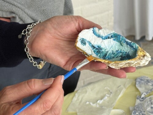 A hand holds an intricately painted oyster shell, with a brush adding gold details to the edges, against a soft background.