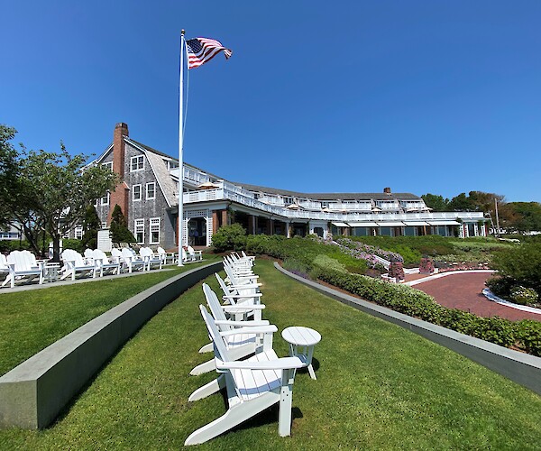 A building with a classic design, white chairs on a lawn, an American flag waving, and a blue sky.