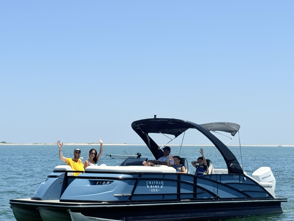 A group of people is waving while on a boat labeled "Chatham Bars Inn" on the water. The sky is clear and blue.