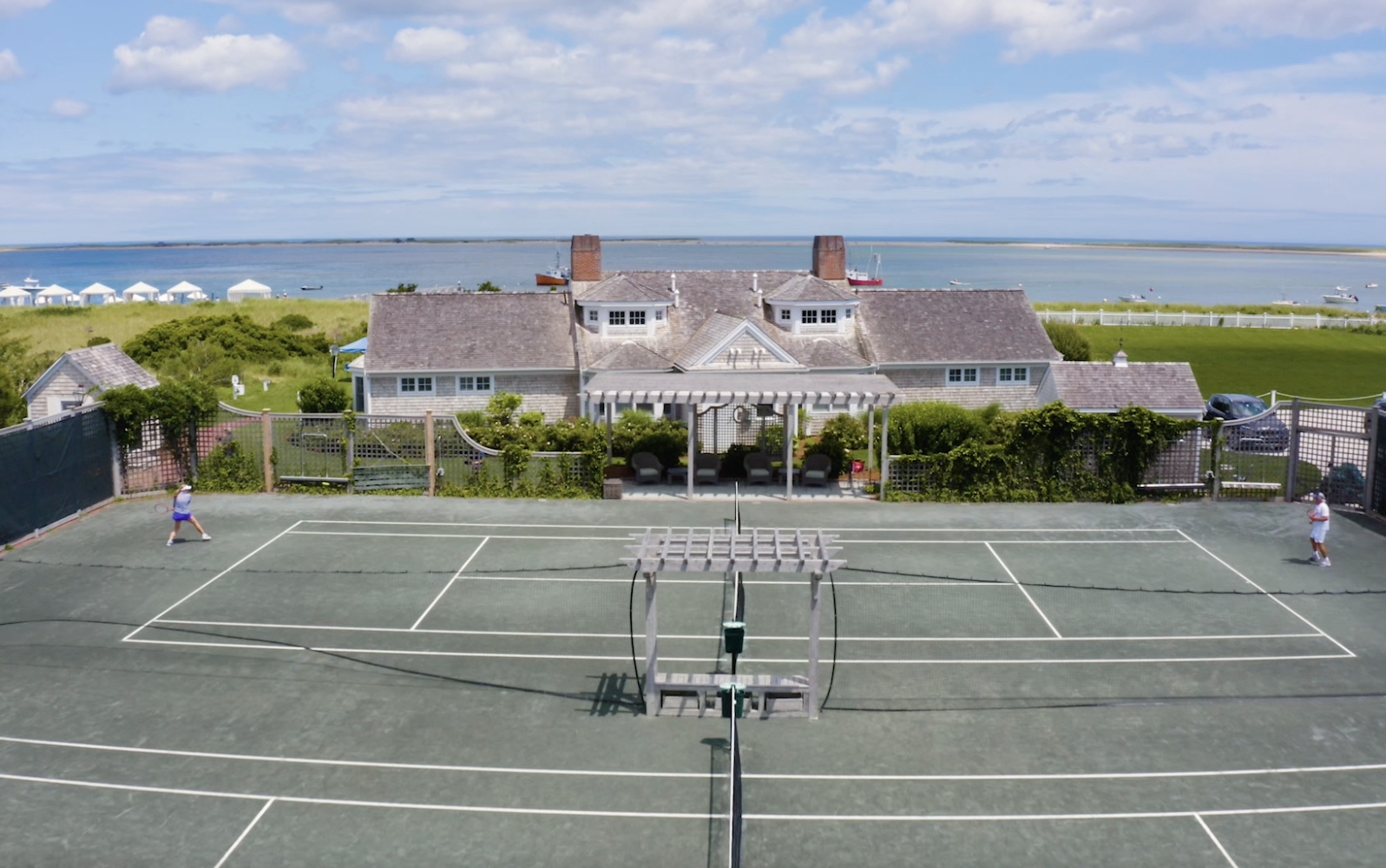 A tennis court with two players is in the foreground, with a house and ocean view in the background under a partly cloudy sky.
