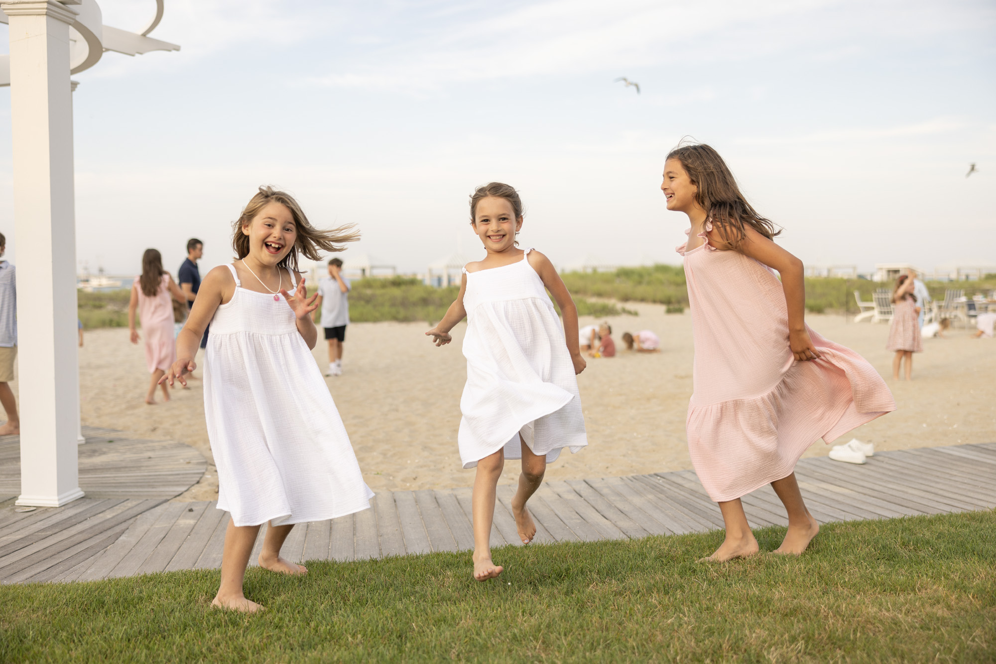 Three children joyfully run on a beach, wearing light dresses, with people relaxing in the background.