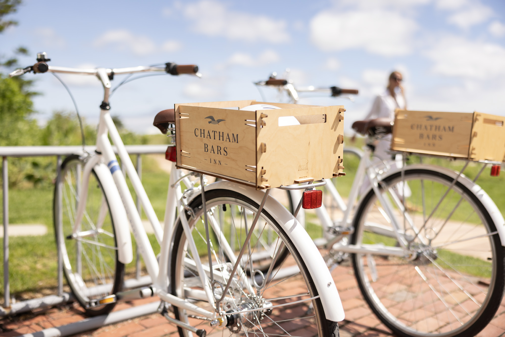 Two white bicycles with wooden boxes labeled "Chatham Bars Inn" are parked outdoors on brick pavement under a partly cloudy sky.