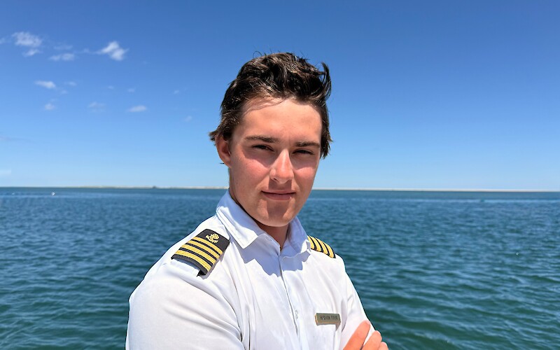 A person is wearing a white uniform with epaulettes, standing by the water under a clear blue sky.