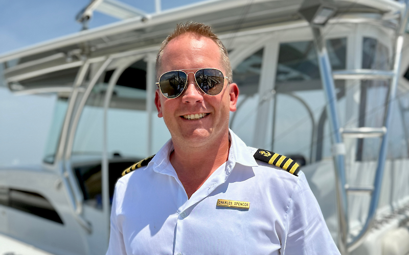 A smiling person in a captain's uniform and sunglasses on a boat deck, with clear skies in the background.