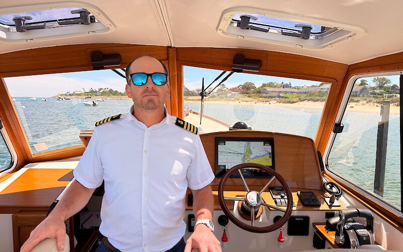 A person in a captain's uniform stands on a boat, with a wooden steering wheel and navigation equipment inside the cabin.