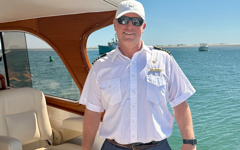A person wearing a white shirt, cap, and sunglasses stands on a boat in front of ocean water under a clear blue sky.