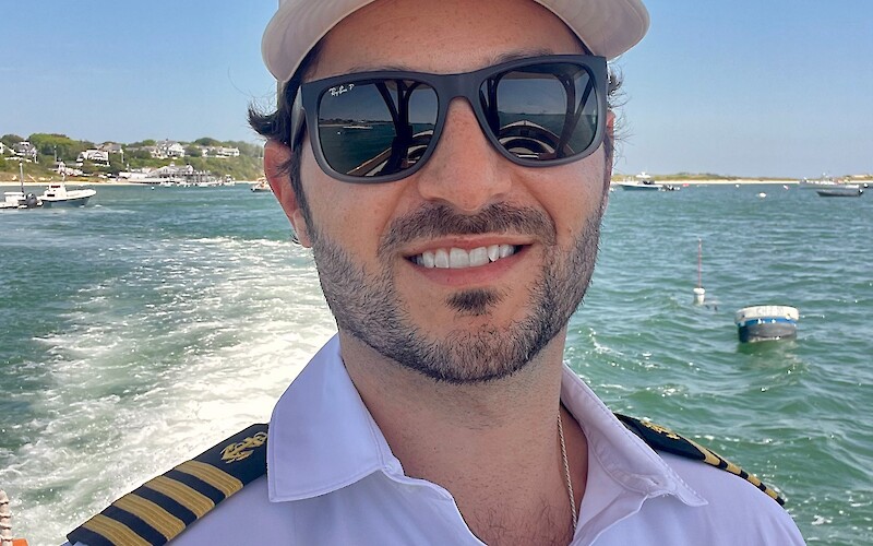 A person wearing a captain's uniform and sunglasses is on a boat with water and a coastline visible in the background.