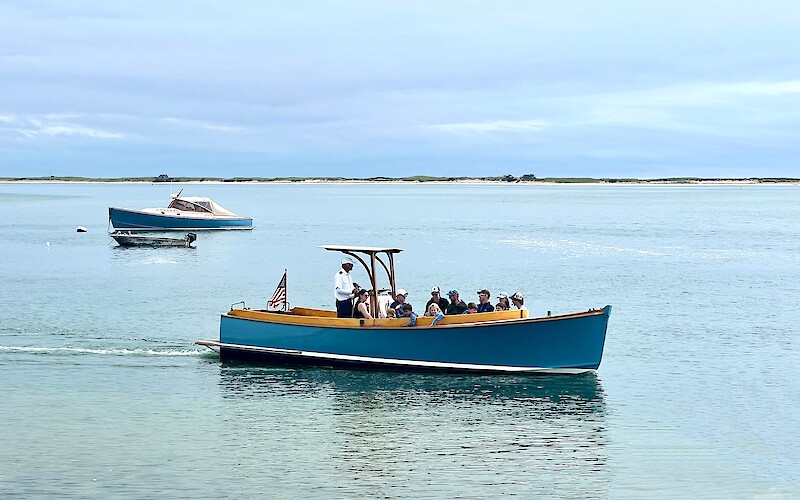 A blue boat with passengers glides on calm water, led by a standing person, while another boat is moored in the background.