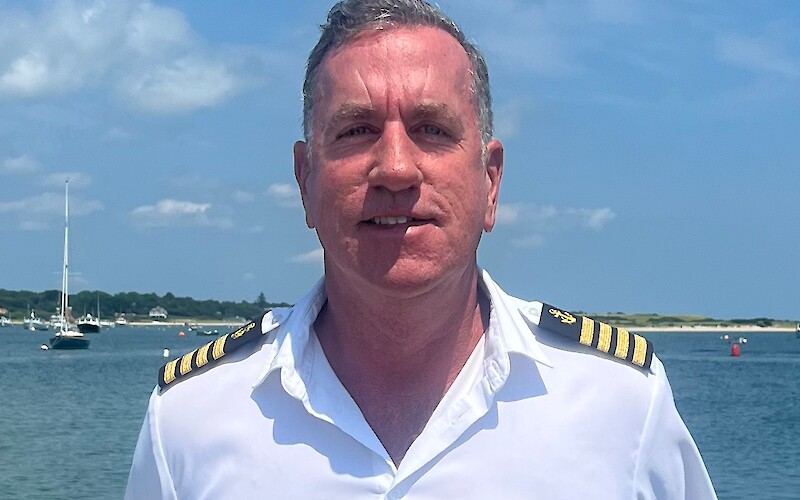 A person in a white uniform with a name tag stands by a body of water, under a blue sky.