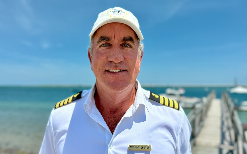 A man in a captain's uniform with a name tag is standing by the sea, with a dock and boats visible in the background.