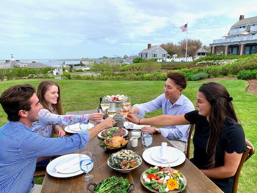 Four people are toasting with drinks at an outdoor table set with food, with a scenic view and American flag in the background.
