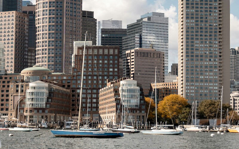 A city skyline with tall modern buildings and a waterfront lined with sailboats under a partly cloudy sky.
