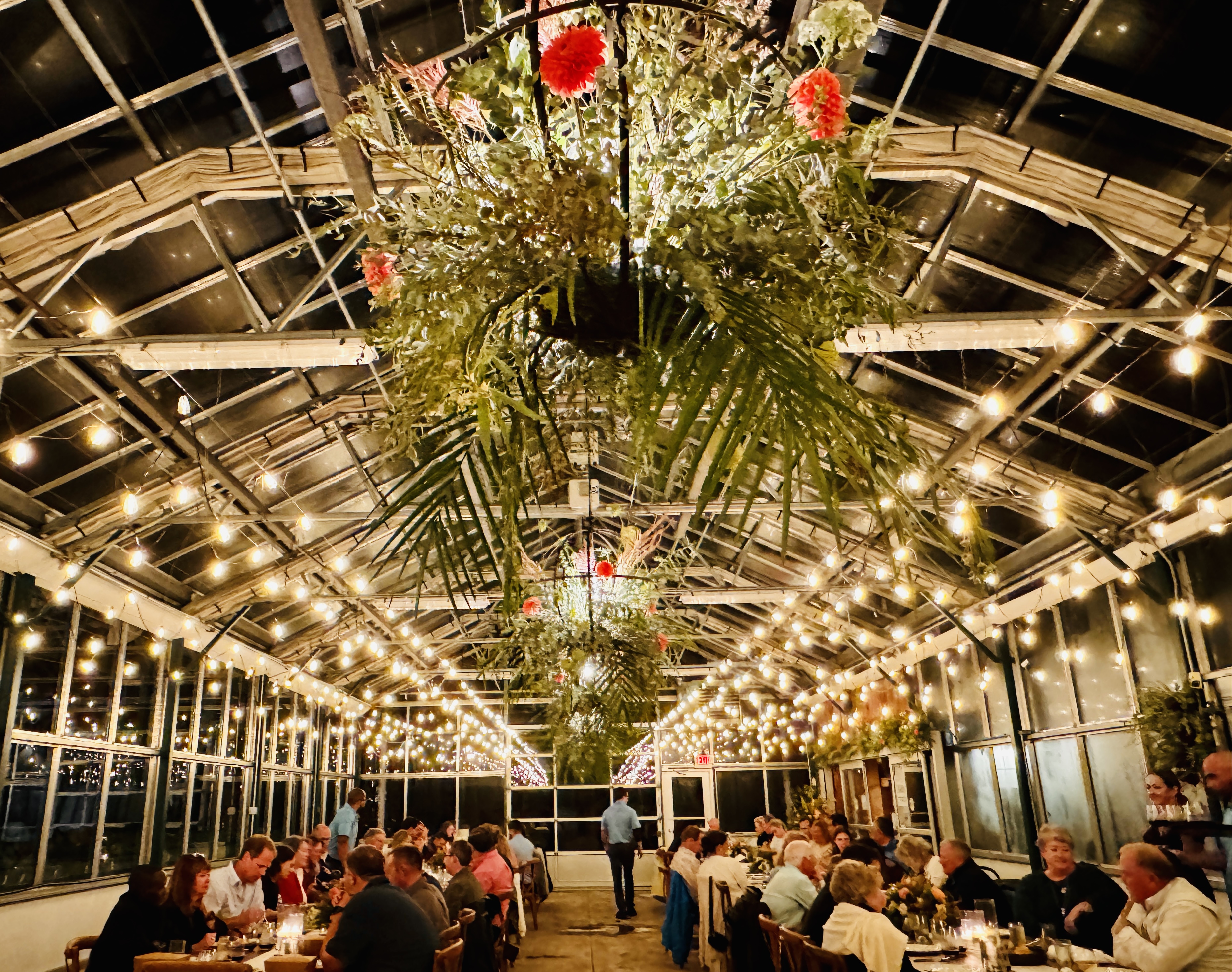 A greenhouse-style setting with string lights, hanging plants, and people dining at long tables within a warmly lit, festive atmosphere.