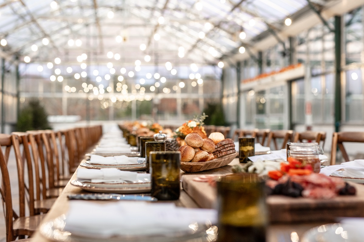 A long, elegantly set dining table with rolls, glasses, plates, and napkins in a well-lit greenhouse-like structure.
