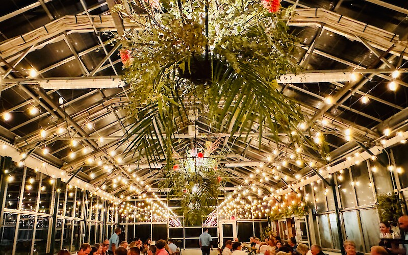 A greenhouse-style venue with tables, people dining, hanging plants, and string lights creating a warm ambiance.