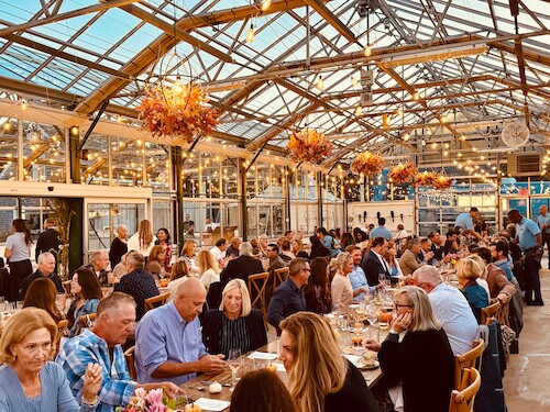 People are enjoying a dinner event in a greenhouse with warm lighting and floral decorations, creating a festive atmosphere.