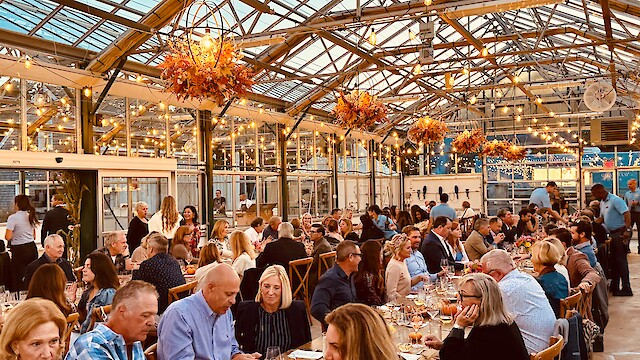 People are enjoying a dinner event in a greenhouse with warm lighting and floral decorations, creating a festive atmosphere.