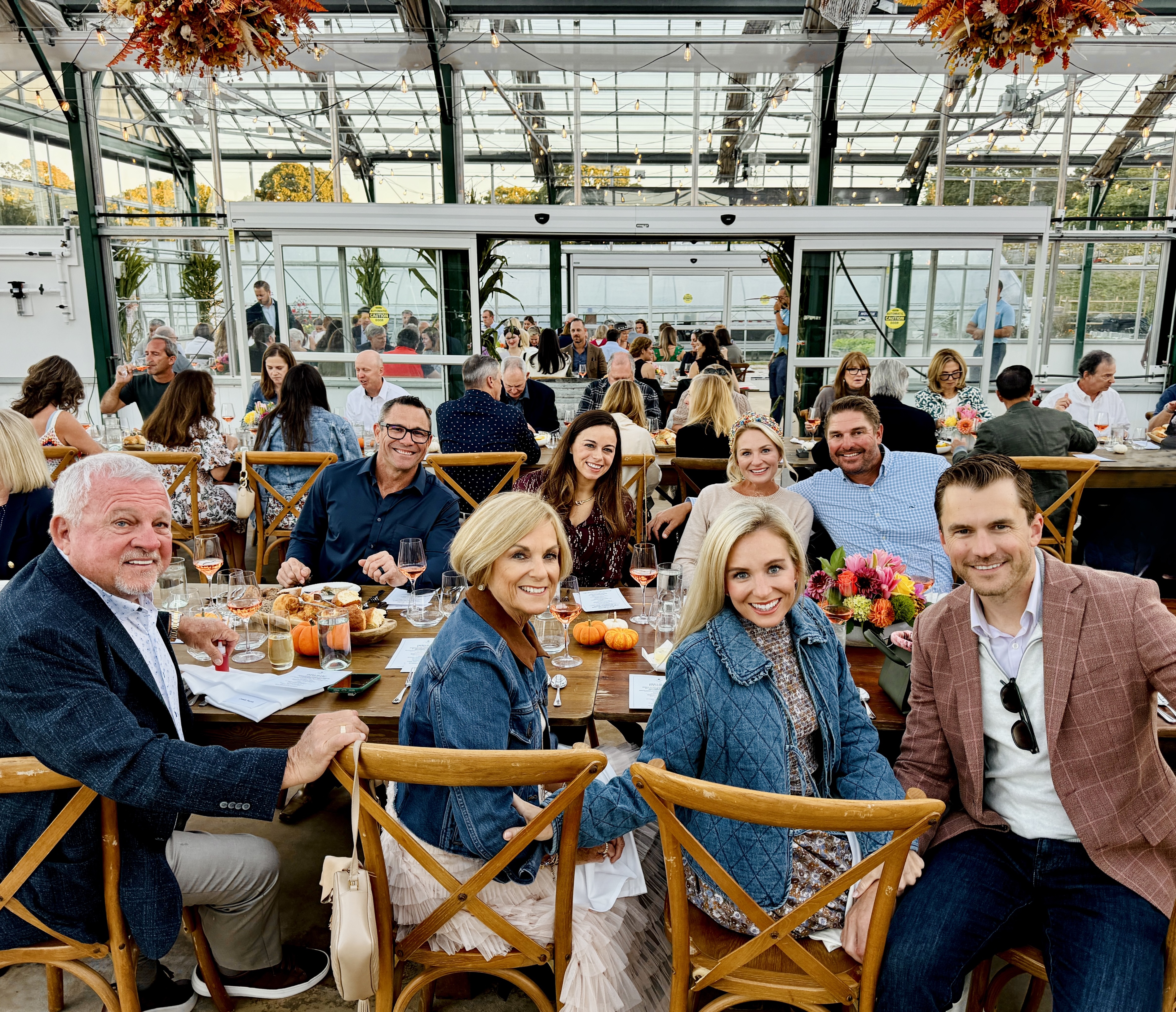 A group of people seated at a long table enjoying a gathering in a greenhouse setting, with autumn decorations hanging above.