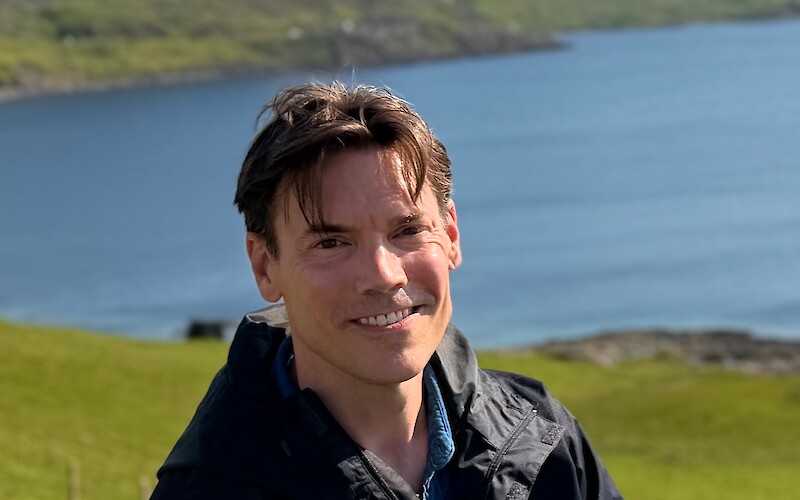 A person smiling, wearing a black jacket, with a scenic backdrop of hills, cliffs, and a body of water under a clear sky.