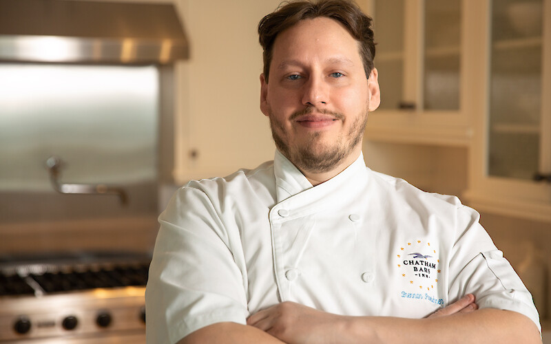 A chef in a white uniform stands confidently with arms crossed in a kitchen setting.