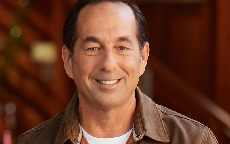 A person wearing a brown jacket and white shirt is smiling, standing indoors with a blurred background.