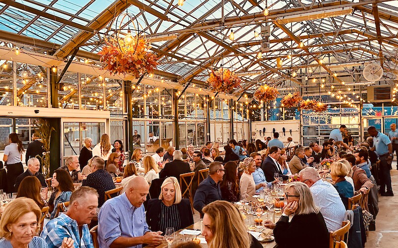 A well-lit greenhouse venue hosts a large dining event with numerous people seated at long tables, surrounded by string lights and decor.