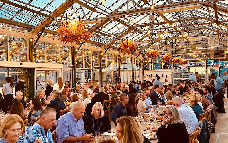 A lively dinner event in a glass-roofed venue with decorative lights and floral arrangements. Guests are seated at long tables.