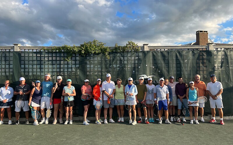 A group of people with tennis gear stands on a tennis court in front of a fence, posing for a photo under a partly cloudy sky.