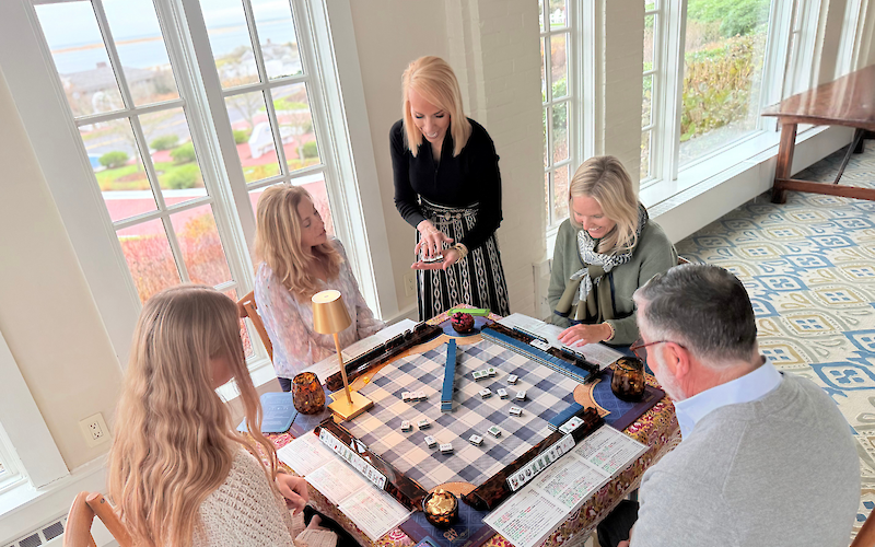 A group playing Mahjong around a table, laughing and enjoying a game, set in a bright room with large windows and scenic views.